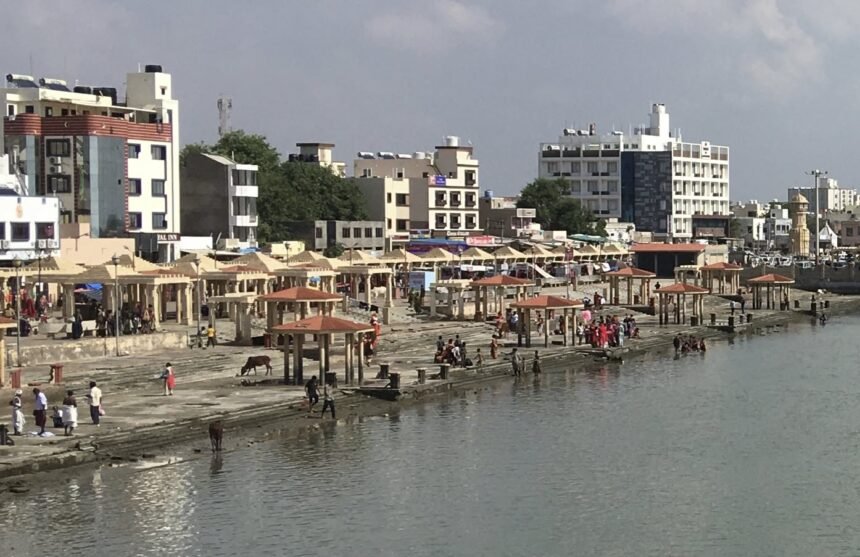 A wide daytime view of Gomti Ghat featuring rows of red-roofed gazebos along the stone steps, with pilgrims bathing in the river, cows on the banks, and city buildings in the background.