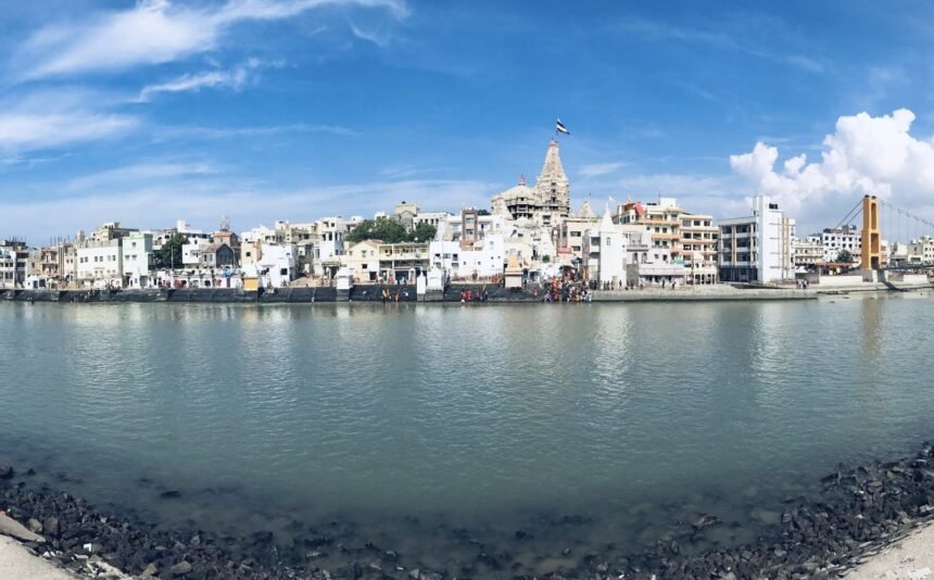 A wide panoramic daytime view of the Gomti River in Dwarka, featuring a rocky foreground, bustling stepped banks, the towering Dwarkadhish Temple spire, and the Sudama Setu bridge under a bright blue sky.