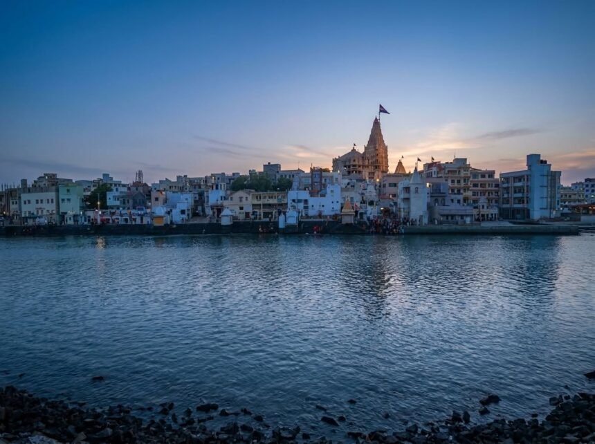 A serene dusk view across the calm waters of Gomti Ghat, featuring the towering spire of the Dwarkadhish Temple and traditional buildings along the bustling riverbanks under a soft twilight sky.