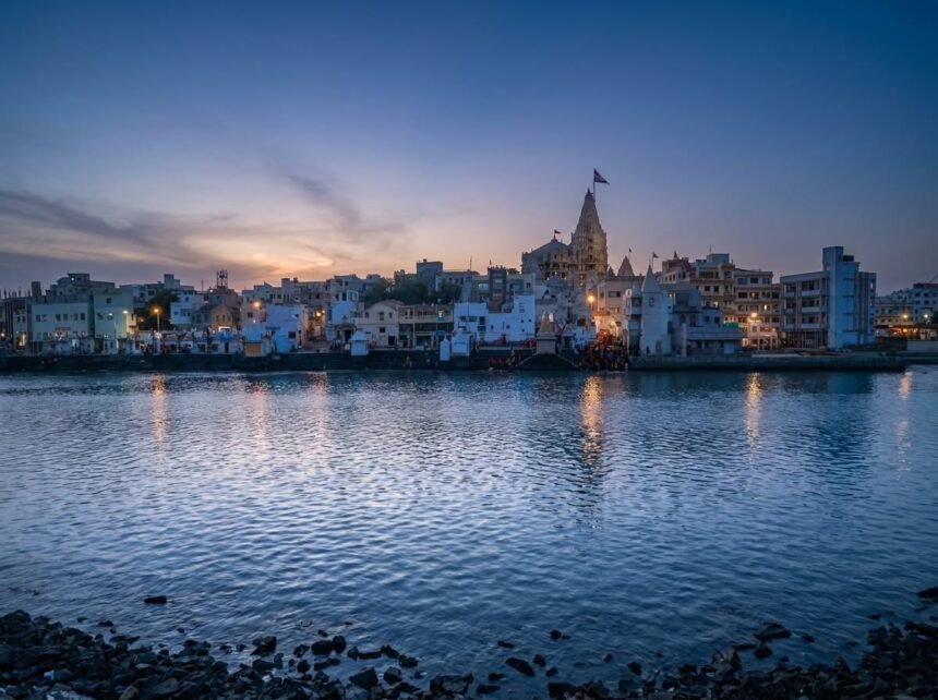 A serene early morning view across the rippling blue waters of Gomti Ghat, showing the Dwarkadhish Temple and city buildings softly illuminated against a dawn sky, with a rocky shoreline in the foreground.