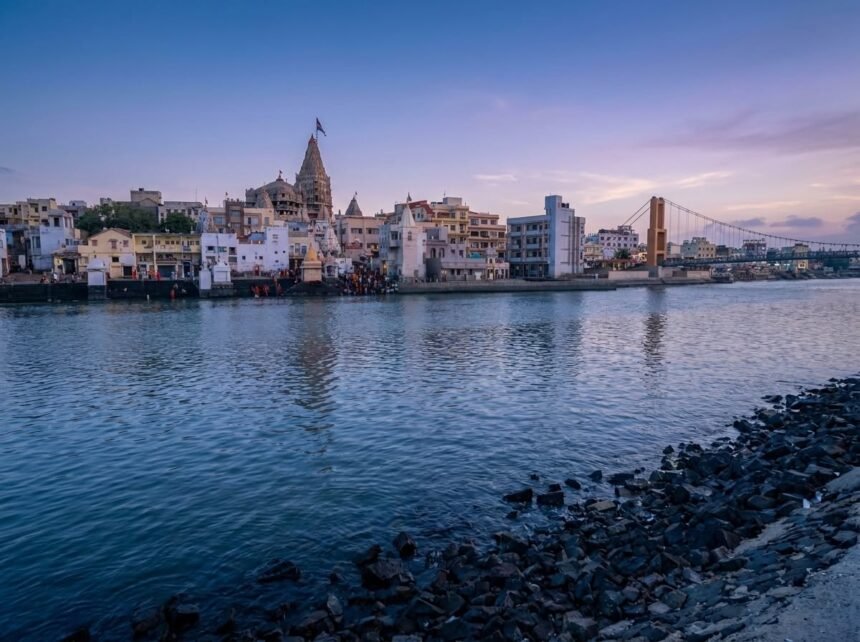 A wide twilight view of Gomti Ghat viewed from a rocky shoreline, showing the Dwarkadhish Temple spire, bustling riverbanks, and the Sudama Setu suspension bridge against a purple and pink early morning sky.