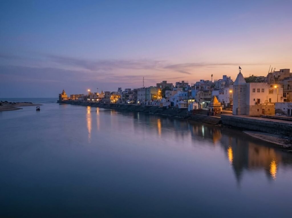 A tranquil evening view of Gomti Ghat featuring illuminated temples and stepped stone banks reflecting in the calm river under a purple and orange sunset sky.