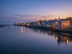 A tranquil evening view of Gomti Ghat featuring illuminated temples and stepped stone banks reflecting in the calm river under a purple and orange sunset sky.