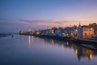 A tranquil evening view of Gomti Ghat featuring illuminated temples and stepped stone banks reflecting in the calm river under a purple and orange sunset sky.
