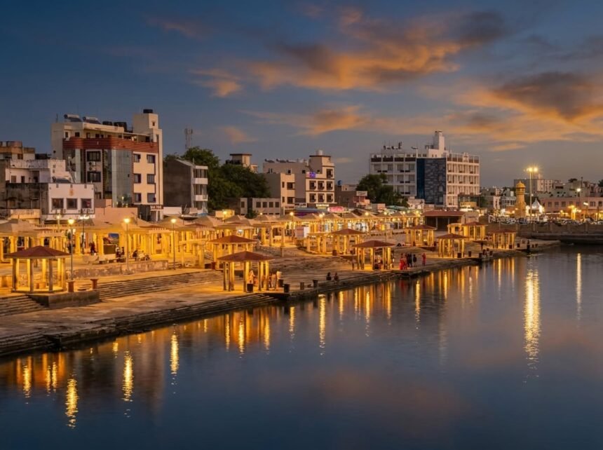 An evening view of the Gomti Ghat riverfront featuring warmly lit pavilions, wide stone steps, and city buildings reflecting in the calm river beneath a striking dark blue sky with vibrant orange sunset clouds.