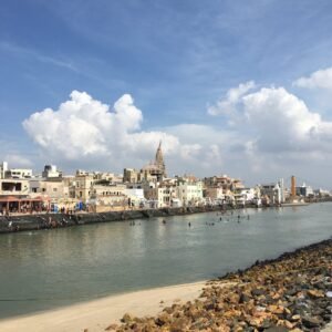 A wide daytime view of Gomti Ghat showing pilgrims bathing in the river, stepped stone banks, and traditional architecture with a prominent temple spire against a blue, cloudy sky.