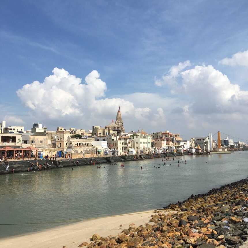 A wide daytime view of Gomti Ghat showing pilgrims bathing in the river, stepped stone banks, and traditional architecture with a prominent temple spire against a blue, cloudy sky.