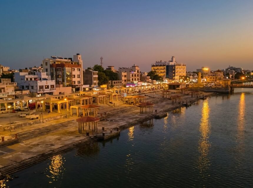 Elevated dusk view of Gomti Ghat's stone promenade featuring warmly lit pavilions and city buildings beside a calm river under a clear twilight sky.