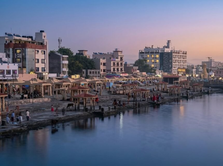 An phenomenal twilight view of Gomti Ghat in Devbhumi Dwarka, featuring traditional pavilions, stone steps, and modern illuminated hotels along the peaceful riverbank.